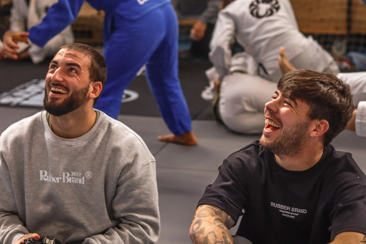 Two men in Rubber Brand Jiu Jitsu apparel smiling on gym mat with others training in background